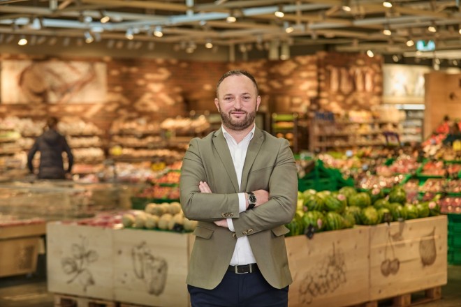 Ein Mann sitzt mit einem Laptop am Tisch und lächelt in die Kamera.