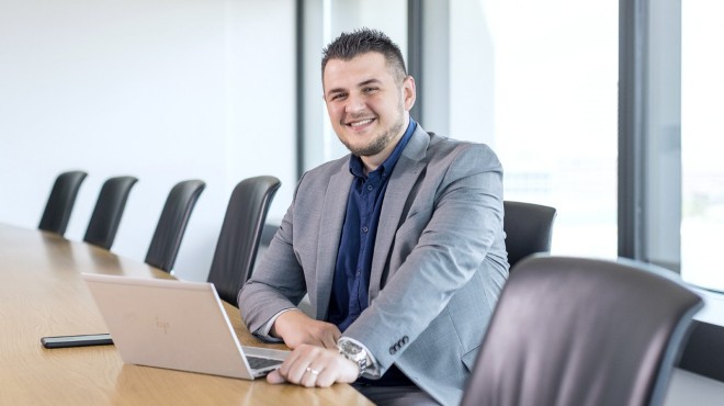 Ein Mann sitzt mit einem Laptop am Tisch und lächelt in die Kamera.