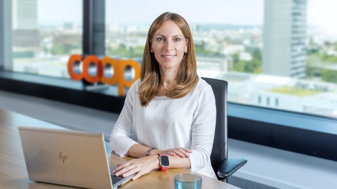 Eine Frau sitzt mit einem Laptop am Tisch und lächelt in die Kamera.