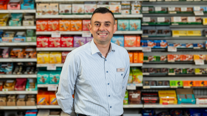 Un uomo sorride all'obiettivo di fronte a uno scaffale di derrate alimentari.